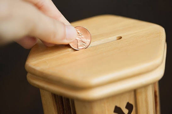 A hand placing a coin into a wooden tzedakah box