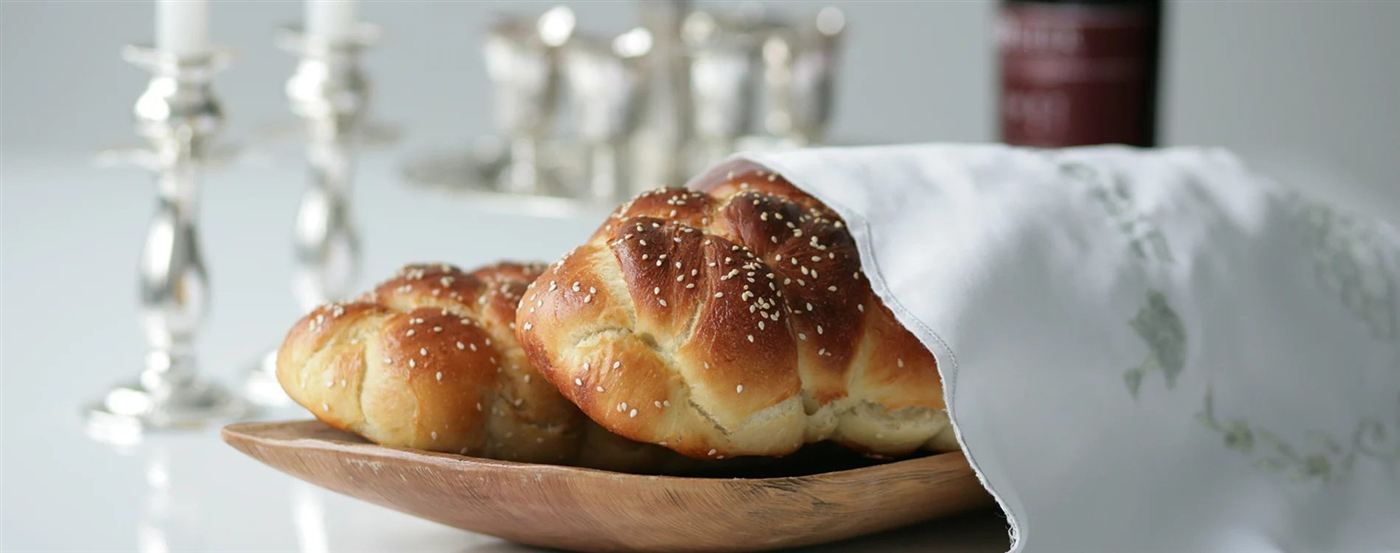 Braided challah loaves wrapped in a cloth beside silver Shabbat candlesticks and kiddush cups